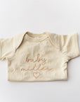 Personalized embroidered onesie featured in natural color with the embroidery in champagne color "Baby Miller and a heart shape" folded on a white background.