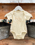 Personalized embroidered onesie featured in natural with the embroidery in champagne color "Baby Miller and a heart shape" hanged on a wooden banister in a white hanger with two gray buckets on each side of it.