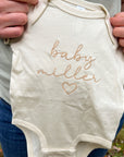 Personalized embroidered onesie featured in natural color with the embroidery in champagne color "Baby Miller and a heart shape" being held by a dad on a nature background.
