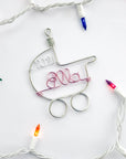 A wire-wrapped baby stroller ornament, featuring the name "Ella" and the year "2021", is nestled among a string of colorful Christmas lights.