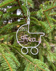 The image shows a wire-wrapped baby stroller ornament, one in silver aluminum featuring the name "Ava" in light pink and the year "1991," all set against a Christmas tree background.