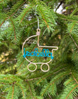 A wire-wrapped baby stroller ornament in gold aluminum, featuring the name "Isabella" in turquoise and the year "2024" in gold, is nestled among a Christmas tree hung by a silver hook.