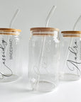 The image features three personalized glass straw tumblers, each with a cylindrical shape, bamboo lid, and glass straw. From left to right, they are personalized with the names and titles: Cassidy, Bridesmaid in black lettering; Sierra, Bride in the center in white lettering; and Andra, Bridesmaid on black lettering on the right, all set against a white background.