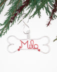 Personalized dog ornament in silver aluminum wire a dog bone outline and the name Mila in red aluminum wire, The background is white that has christmas tree branches.