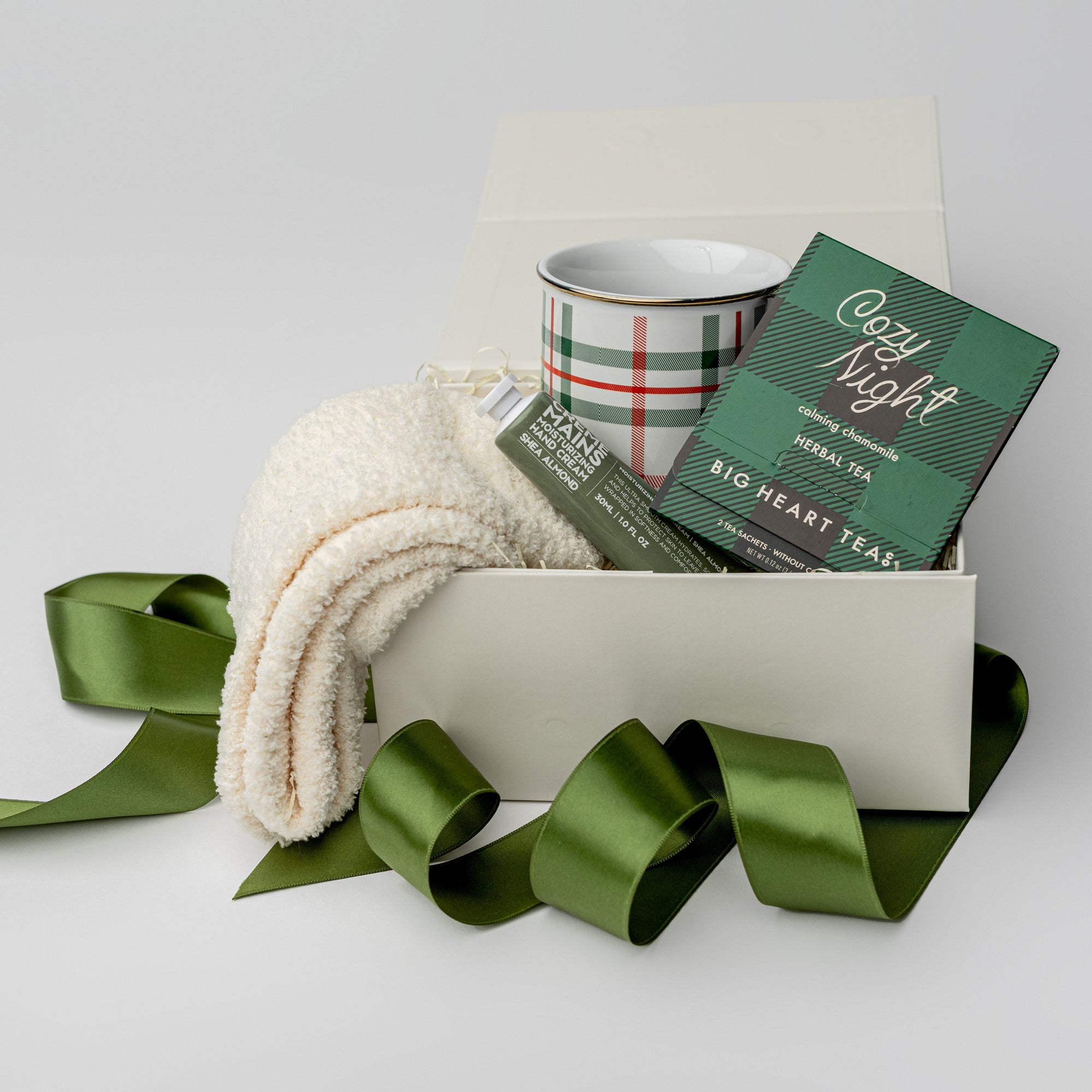 Petite Cozy at Home Holiday Gift is featured in a white box wrapped with a green ribbon. Inside the box, on the right, are white cozy socks; in the middle, a plaid mug with green and red lines; and on the left, a Cozy Night herbal tea in black and green plaid packaging. Everything is displayed on a white background.