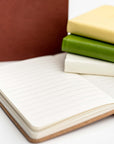 Collection of pocket soft cover notebooks in the different colors they come burgundy, rust, yellow, green and cream. The rust is opened showing the inside of the notebook white pages with black lines.
