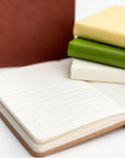 Collection of pocket soft cover notebooks in the different colors they come burgundy, rust, yellow, green and cream. The rust is opened showing the inside of the notebook white pages with black lines.