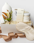 A white gift box with ivory paper shred and tied with a brown ribbon on a white background. Inside the box are a stone oil diffuser, a bottle of Vitruvi organic eucalyptus essential oil, Herbivore Botanicals coconut milk bath soak, an ivory satin sleep mask, Formulary 55 eucalyptus & peppermint sea mineral foot soak, a MAST organic almond dark chocolate bar, and a handmade dried floral mini bouquet — a soothing self-care gift box.