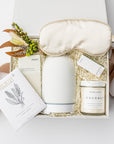 A white gift box with ivory paper shred and tied with a brown ribbon on a white background. Inside the box are a stone oil diffuser, a bottle of Vitruvi organic eucalyptus essential oil, Herbivore Botanicals coconut milk bath soak, an ivory satin sleep mask, Formulary 55 eucalyptus & peppermint sea mineral foot soak, a MAST organic almond dark chocolate bar, and a handmade dried floral mini bouquet — a soothing self-care gift box.