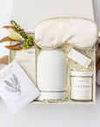 A white gift box with ivory paper shred and tied with a brown ribbon on a white background. Inside the box are a stone oil diffuser, a bottle of Vitruvi organic eucalyptus essential oil, Herbivore Botanicals coconut milk bath soak, an ivory satin sleep mask, Formulary 55 eucalyptus & peppermint sea mineral foot soak, a MAST organic almond dark chocolate bar, and a handmade dried floral mini bouquet — a soothing self-care gift box.