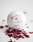 Rose Petal Bath Bomb, featured in its white transparent wrapping with a label that includes a rose illustration, pink details, and black lettering, surrounded by rose petals on a white background.