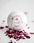 Rose Petal Bath Bomb, featured in its white transparent wrapping with a label that includes a rose illustration, pink details, and black lettering, surrounded by rose petals on a white background.