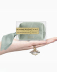 A hand holds a clear box containing a sage green velvet emergency kit for bridesmaids, with a matching sage green ribbon and a gold label. A gold ring is draped over the hand against a clean white background.