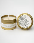 Santal and vanilla travel size candle featured in its gold tin and white label with flower illustration in black on lid and black lettering on tin on a white background.