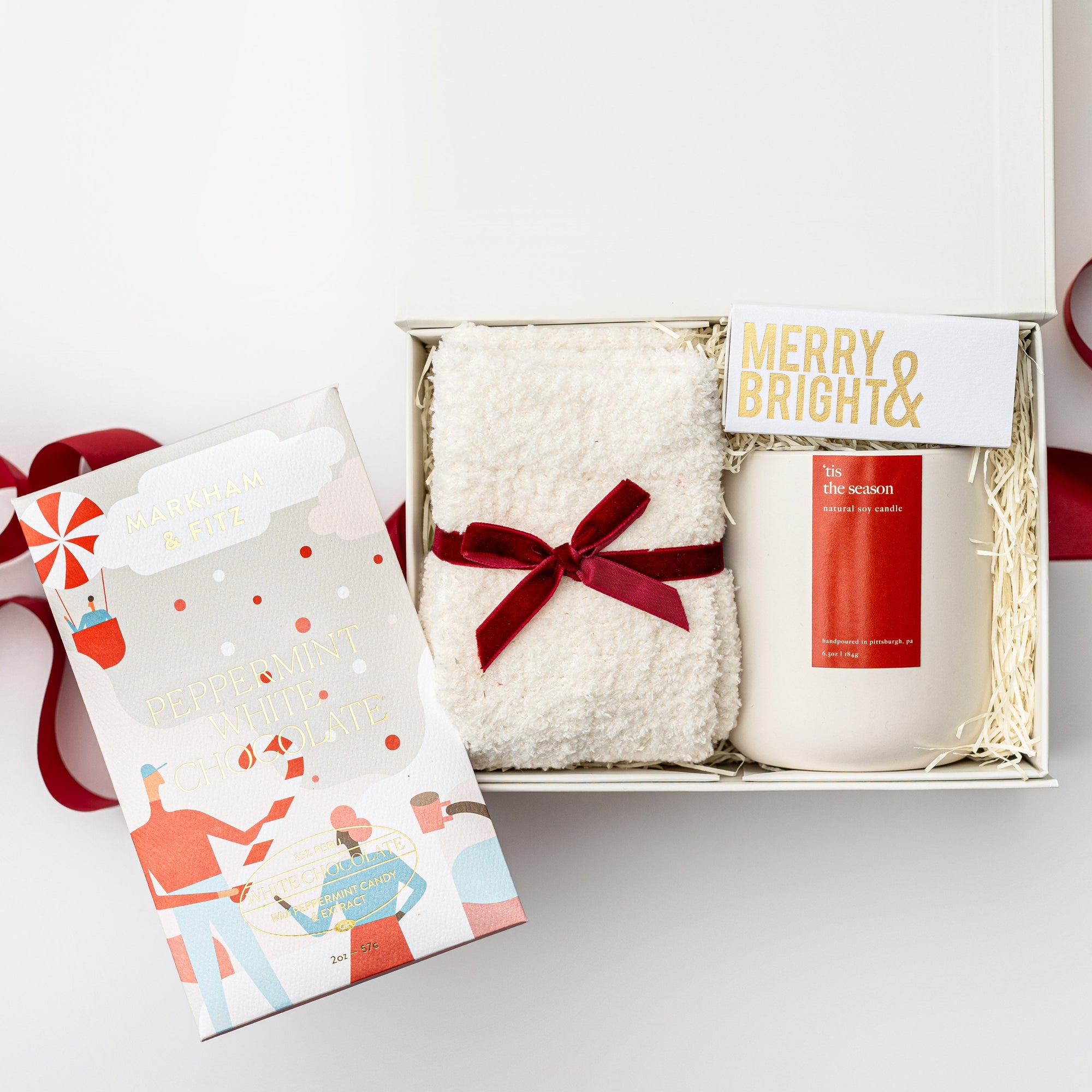 Secret Santa holiday gift box is featured in small white box with white shred with red ribbon underneath the box  outside the box on its right side peppermint chocolate bar with Christmas graphic, inside on top in the left white cozy socks wrapped in a red ribbon. On the right merry & bright gold foil matchbox and a white ceramic  tis the season candle with red label, all on a white background.