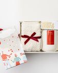 Secret Santa holiday gift box is featured in small white box with white shred with red ribbon underneath the box  outside the box on its right side peppermint chocolate bar with Christmas graphic, inside on top in the left white cozy socks wrapped in a red ribbon. On the right merry & bright gold foil matchbox and a white ceramic  tis the season candle with red label, all on a white background.