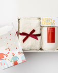 Secret Santa holiday gift box is featured in small white box with white shred with red ribbon underneath the box outside the box on its right side peppermint chocolate bar with Christmas graphic, inside on top in the left white cozy socks wrapped in a red ribbon. On the right merry & bright gold foil matchbox and a white ceramic tis the season candle with red label, all on a white background.