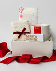 Secret Santa holiday gift box is featured a white small box with a red ribbon around it. inside of the box white on the left cozy white sock tied in red ribbon on the right this is the season candle with red label in the back peppermint  white chocolate bar and in the front merry & bright gold foil matchbox. All on a white background.