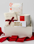 Secret Santa holiday gift box is featured a white small box with a red ribbon around it. inside of the box white on the left cozy white sock tied in red ribbon on the right this is the season candle with red label in the back peppermint white chocolate bar and in the front merry & bright gold foil matchbox. All on a white background.