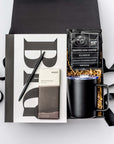 A top view of the Seize the Day Gift Box presented in a black gift box filled with brown shred and tied with a black ribbon, featuring a Big Idea sketchbook, a minimalist black bullet pen, a 12 oz. black vacuum insulated camp mug, a Copper Cow single-serve pour-over coffee, and a MAST organic dark chocolate bar (80% cacao), all arranged on a white background.