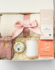 Top view of the “Sending Love” gift box displayed in a white box with a pink ribbon and ivory shred, containing a soft knit ivory throw blanket, a 12oz speckled ceramic mug, Cup of Love rose tea for two, a creamy milk chocolate bar, heart cozy socks, and a 4oz hand-poured Sweet Fig candle.
