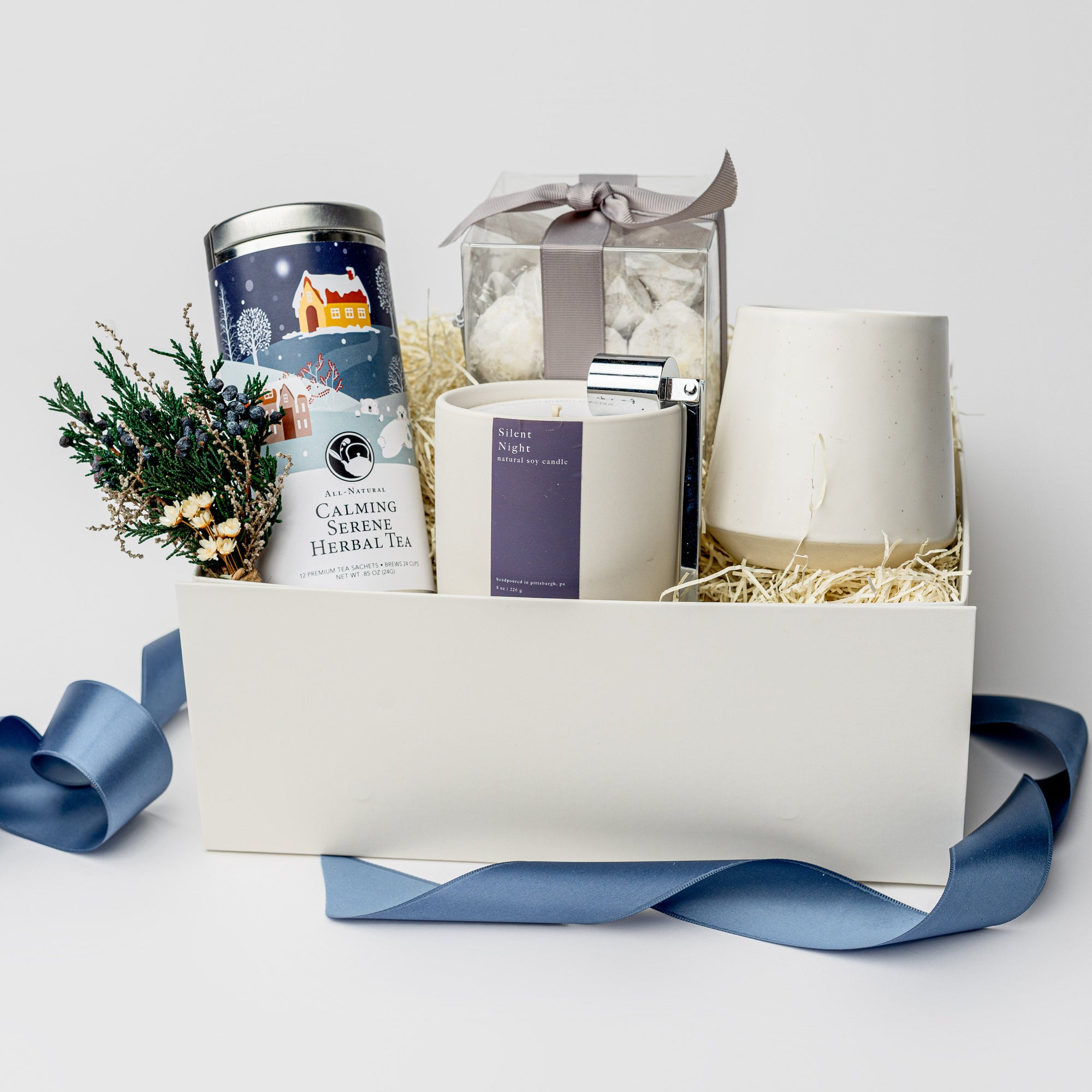 Silent Night Holiday Gift Box is featured open, revealing a white gift box with a blue ribbon in front of it. All arranged in front of white shred on the left side floral bundle, calming serene herbal tea, in the middle tea cakes, silent night candle with silver snuffer on top and on the left white speckled ceramic mug,. All on a white background.