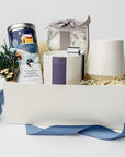 Silent Night Holiday Gift Box is featured open, revealing a white gift box with a blue ribbon in front of it. All arranged in front of white shred on the left side floral bundle, calming serene herbal tea, in the middle tea cakes, silent night candle with silver snuffer on top and on the left white speckled ceramic mug,. All on a white background.