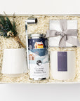 Silent Night Holiday Gift Box is featured open, a white gift box with a blue ribbon crossing underneath. Inside the box, arranged on top of white shred. On the left side, a floral bundle placed on top of a speckled white cup. In the middle, a silver candle snuffer and a calming serene herbal tea in a round silver tin with a Christmas-themed graphic label. On the right, a clear box containing tea cakes tied with a silver ribbon and a white ceramic “Silent Night” candle with a purple label, white background.