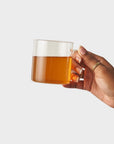 A hand holding a clear glass mug filled with amber tea against a plain light gray background.