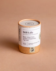 Still life candle is featured in its cylindrical container with tan bottom and rim white label black lettering on a beige background.