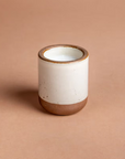 Still life candle is featured in its white ceramic jar with tan bottom and rim in a beige background.