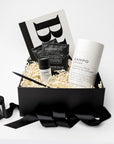 A black gift box filled with spa and self-care items, including a white and black CAMPO diffuser, a small black bottle, two black CAMPO Pour Over Coffee sachets, and a black pen, arranged in white crinkle paper with a black ribbon in front on a white background.