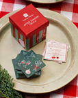 A boxed card game called Talking Tossers After Dinner Trivia sits on a beige ceramic plate over a red-and-white checkered tablecloth. The red box has green stripes and a small illustration.
