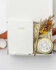 A top view of the Thankful For You Gift Box presented in a white gift box filled with ivory shred and tied with a toffee-colored ribbon, featuring an ivory softcover pocket journal, a minimalist gold bullet pen, a 4 oz. hand-poured White Tea candle, and a handmade dried floral mini bouquet, all arranged on a white background.