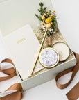 A top view of the Thankful For You Gift Box presented in a white gift box filled with ivory shred and tied with a toffee-colored ribbon, featuring an ivory softcover pocket journal, a minimalist gold bullet pen, a 4 oz. hand-poured White Tea candle, and a handmade dried floral mini bouquet, all arranged on a white background.