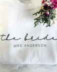 The bride custom name sweatshirt is featured in white color and the personalization in black lettering the bride Mrs. Anderson on a marble like background and a flower on upper right corner.