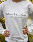 The bride custom name sweatshirt in white personalized in black lettering the bride Mrs. Anderson worn by the bride ina nature background.