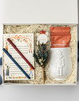 The Nutcracker Holiday Gift Box is featured open, revealing a white gift box with a blue ribbon crossing underneath. Inside the box, all arranged on top of white shred. On the left side, there is a Nutcracker notepad with a blue and red pen on top. In the center, a clear box of candy caramels with a white label, gold rim, and red ribbon sits beside a floral bundle. On the right, there is a white embossed Nutcracker mug containing two orange monstruck chocolate packages, all on a white background.