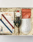 The Nutcracker Holiday Gift Box is featured open, revealing a white gift box with a blue ribbon crossing underneath. Inside the box, all arranged on top of white shred. On the left side, there is a Nutcracker notepad with a blue and red pen on top. In the center, a clear box of candy caramels with a white label, gold rim, and red ribbon sits beside a floral bundle. On the right, there is a white embossed Nutcracker mug containing two orange monstruck chocolate packages, all on a white background.