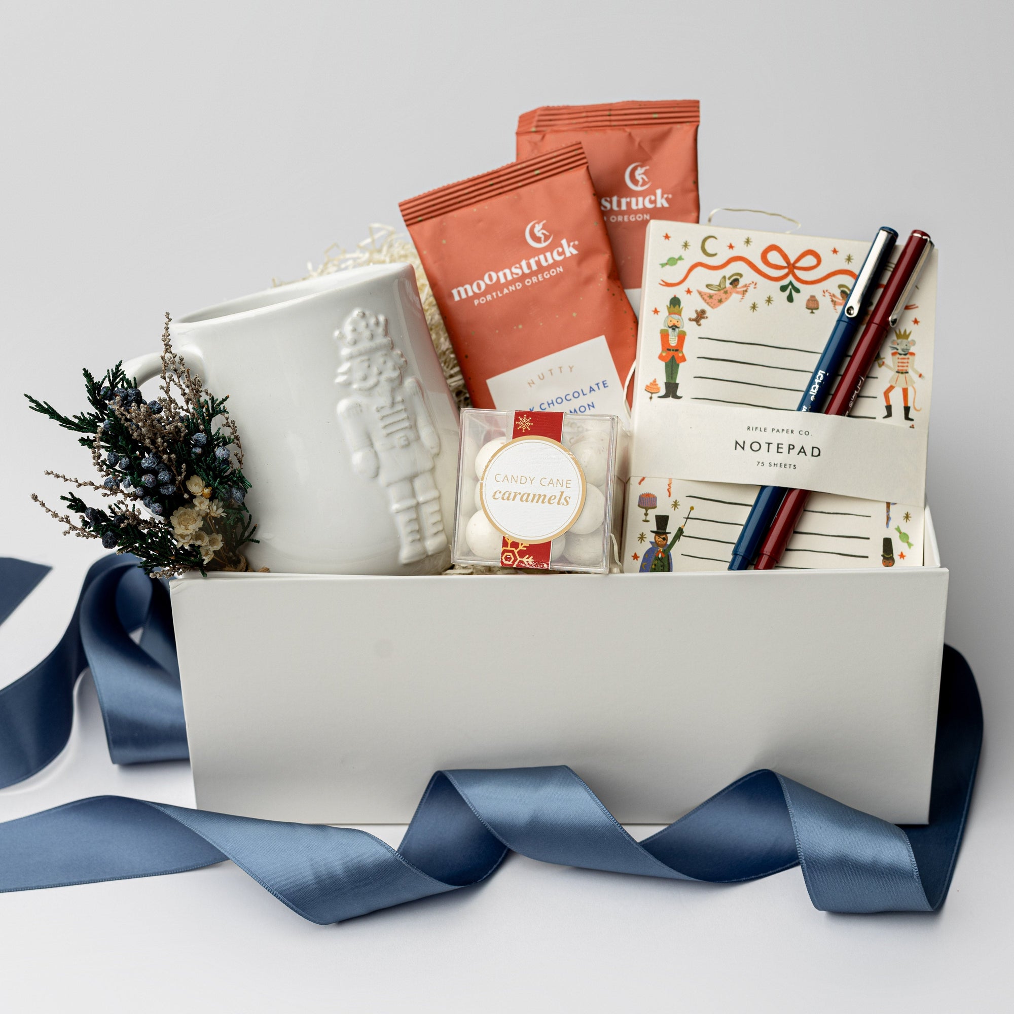 The Nutcracker Holiday Gift Box is featured open, revealing a white gift box with a blue ribbon in front of it. Inside the box, all arranged in front of a white shred. On the left side, there is a white embossed Nutcracker mug beside a floral bundle.  In the center, a clear box of candy caramels with a white label, gold rim, and red ribbon on top of it two orange monstruck chocolate packages . On the right, Nutcracker notepad with a blue and red pen on top , all on a white background.