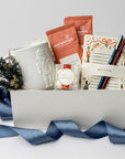The Nutcracker Holiday Gift Box is featured open, revealing a white gift box with a blue ribbon in front of it. Inside the box, all arranged in front of a white shred. On the left side, there is a white embossed Nutcracker mug beside a floral bundle.  In the center, a clear box of candy caramels with a white label, gold rim, and red ribbon on top of it two orange monstruck chocolate packages . On the right, Nutcracker notepad with a blue and red pen on top , all on a white background.