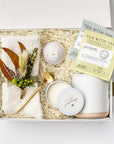 Warmest Wishes sympathy Gift Box presented in a white gift box filled with ivory shred and tied with a green ribbon, featuring a Milk & Honey bath bomb, a 12 oz. speckled ceramic mug, two jasmine green and peppermint tea sachets, a gold stir spoon, ivory plush cloud socks, a 4 oz. hand-poured “No Words, Just Hugs” candle, and a handmade dried floral mini bouquet, all arranged on a white background.