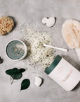 A flat-lay of a skincare product set on a textured gray background, featuring a white jar with a dark green lid labeled 'The Clerody' and 'The Clerydy' cream, alongside a small bowl of green scrub, loose powder, a sponge, a brush, and stones.