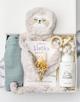 A top view of a baby gift set presented in a white gift box filled with ivory shred, featuring a Slumberkins Slumber Sloth, a muslin cotton swaddle blanket, Night Night baby wash, a silicone and wood baby teether, and a handcrafted dried floral mini bouquet, showing a silver ribbon, all arranged on a white background.