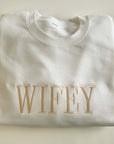 White crewneck sweatshirt featuring “wifey” embroidered in gold lettering, displayed on a white background.