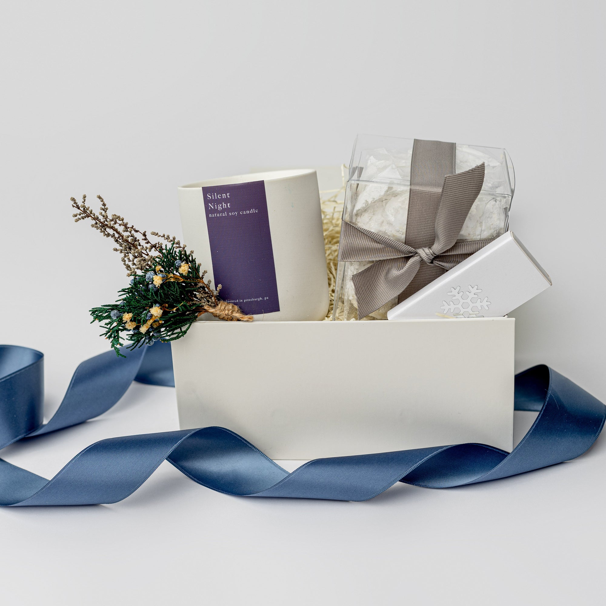 Winter Wonderland Gift Box featured with a white box containing a white ceramic “Silent Night” candle with a purple label and white lettering, a handmade floral mini bundle with blue, yellow, green, and beige flowers wrapped in a twig, a box of tea cakes in a transparent square container tied with a silver ribbon, and a snowflake silver foil candle matchbox. The box is filled with ivory shreds in the back and finished with a blue ribbon in the front, displayed on a white background.