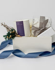 Winter Wonderland Gift Box featured with a white box containing a white ceramic “Silent Night” candle with a purple label and white lettering, a handmade floral mini bundle with blue, yellow, green, and beige flowers wrapped in a twig, a box of tea cakes in a transparent square container tied with a silver ribbon, and a snowflake silver foil candle matchbox. The box is filled with ivory shreds in the back and finished with a blue ribbon in the front, displayed on a white background.