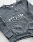 Custom embroidered youth sweatshirt charcoal grey with white “ELIJAH” text on a marble background.