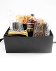 Black box with a set of two crystal tumblers, a double sided golden jigger, bottle of old fashioned syrup with black and gold label, a pack of playing cards with elaborate gold and bronze foil design, pack of dried oranges.