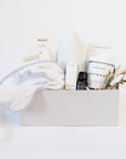 Light grey box containing an Ivory sleep mask, Formulary 55 Eucalyptus & Peppermint foot soak, Mast tea chocolate bar, Vitruvi white stone diffuser, black bottle of Vitruvi eucalyptus oil next to it's white box, glass jar of Herbivore coconut bath soak next to it's white box, dried floral bundle.